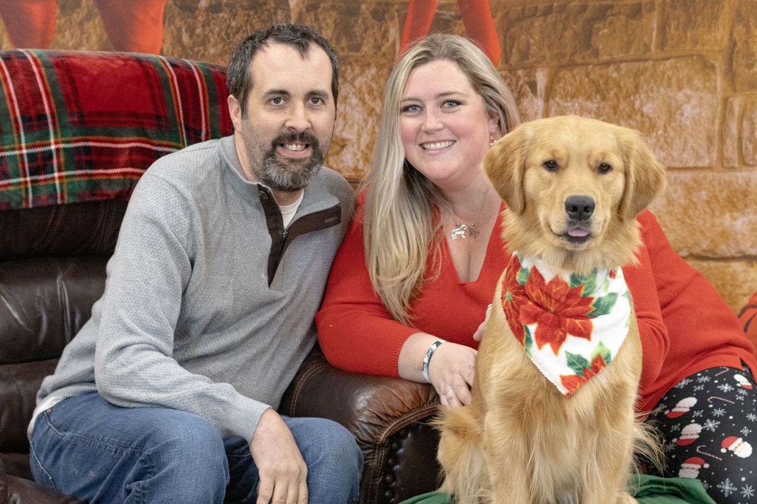 A couple smiles with their golden retriever wearing a festive Christmas bandana for a holiday portrait.