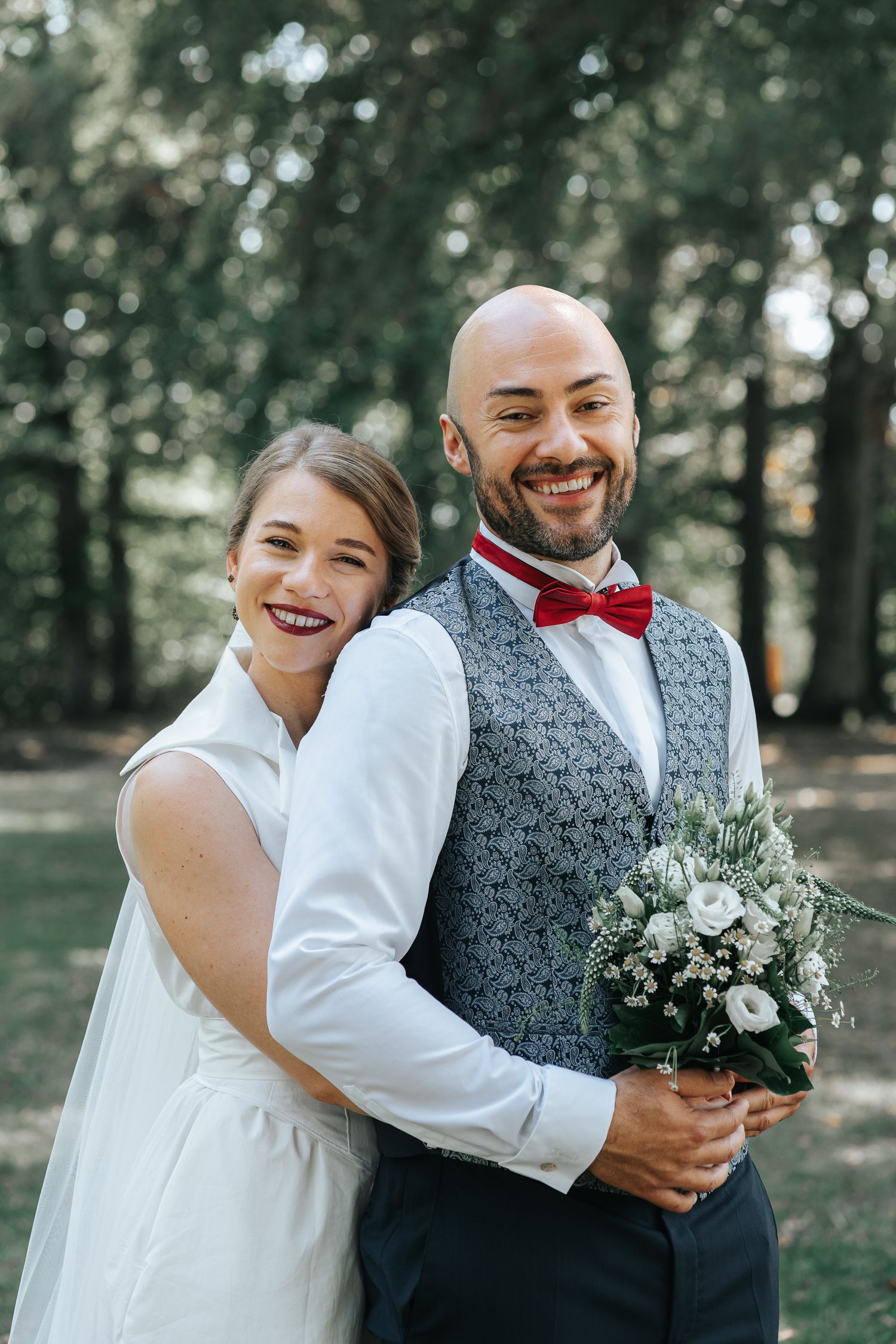 A joyful wedding portrait showing a couple with the groom in a grey vest and red bowtie holding white flowers.
