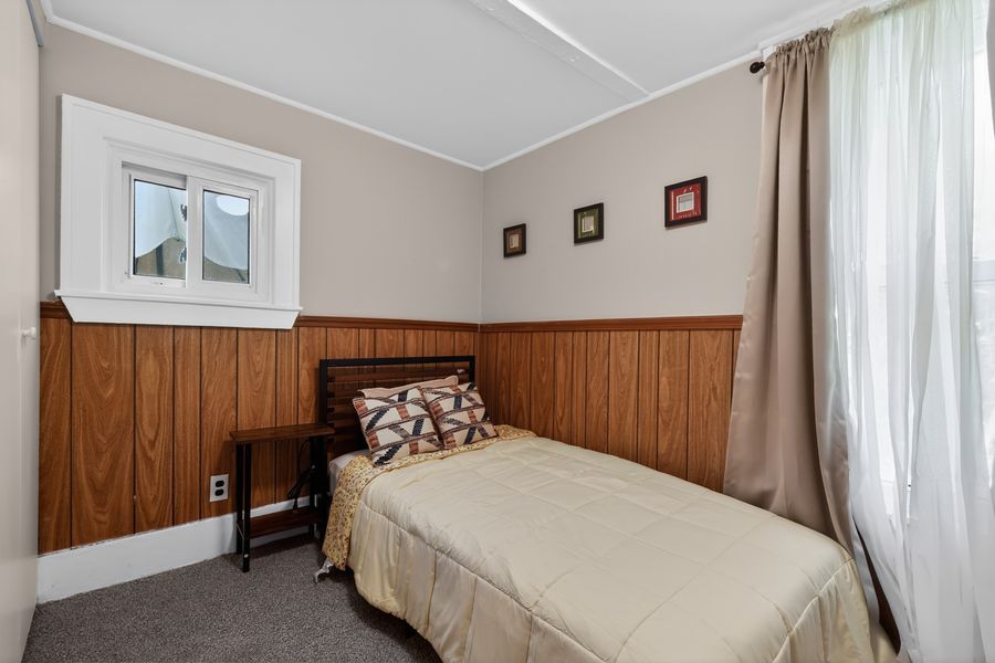 Cozy bedroom with wood panel wainscoting, single bed with neutral bedding, gray carpet, and sheer curtains by the window.