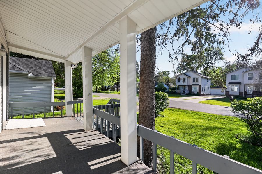 Covered front porch with white railings overlooking a lush green lawn and neighboring homes on a sunny day.