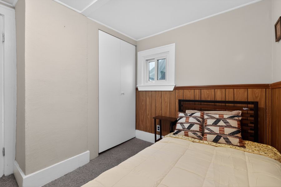 Cozy bedroom with wood-paneled walls, gray carpet, white closet door, and a bed with patterned pillows near a small window.