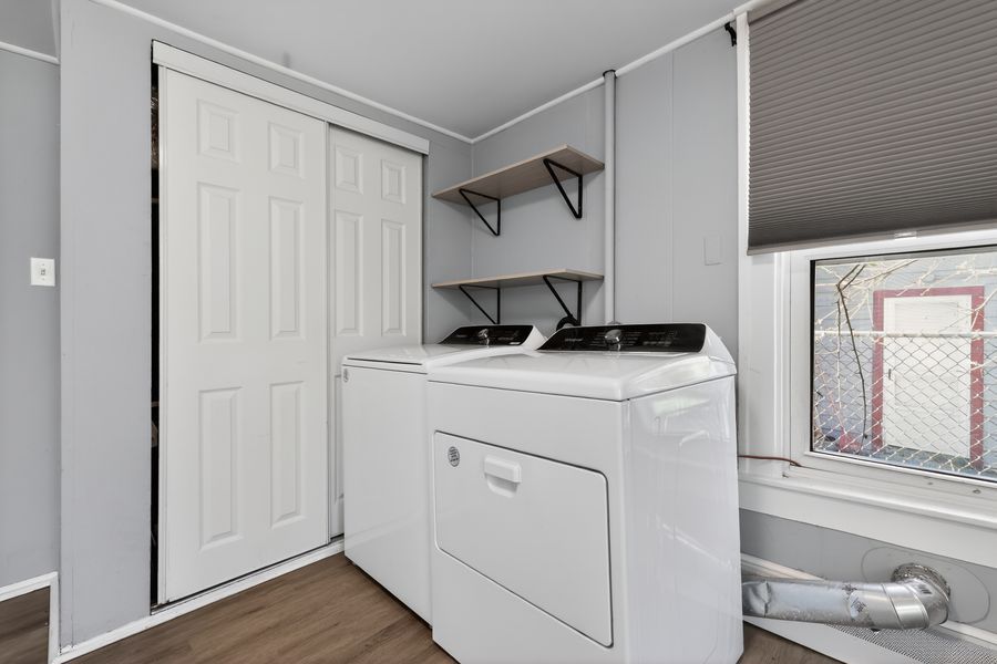 Laundry room with white washer and dryer, wooden shelves, closet door, and hardwood floors.