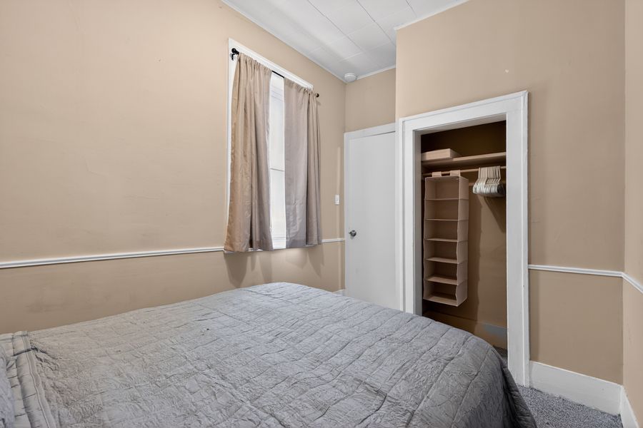 Small bedroom with gray bedding, beige walls, a curtained window, and an open closet with built-in shelving.