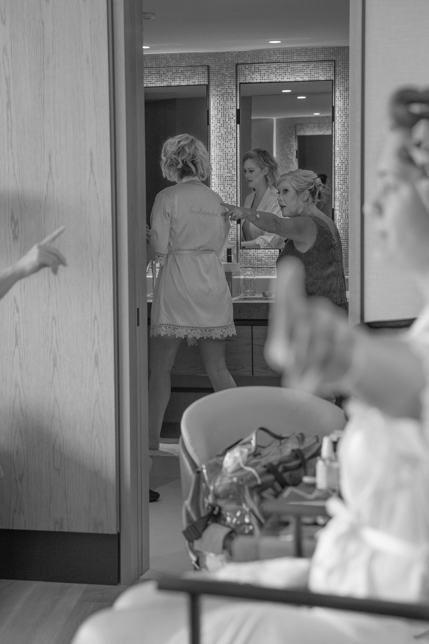 Black and white photo of bridesmaids helping bride get ready in a bathroom mirror reflection.