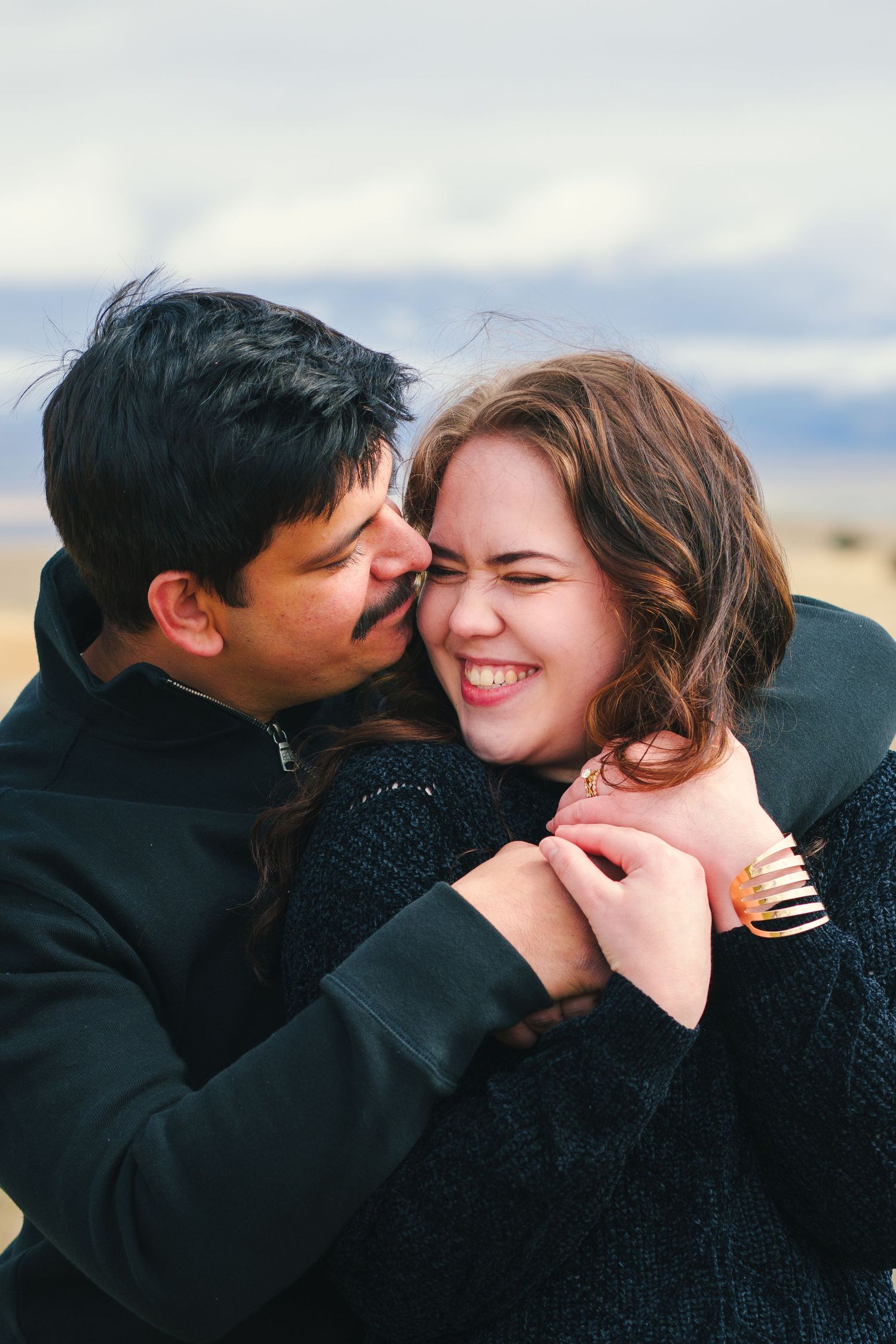 Joyful young couple sharing a playful kiss and laugh in an outdoor winter landscape setting.