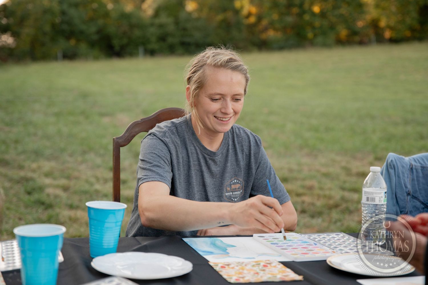 Woman smiling while playing bingo at an outdoor table with blue cups and a water bottle on a black tablecloth.