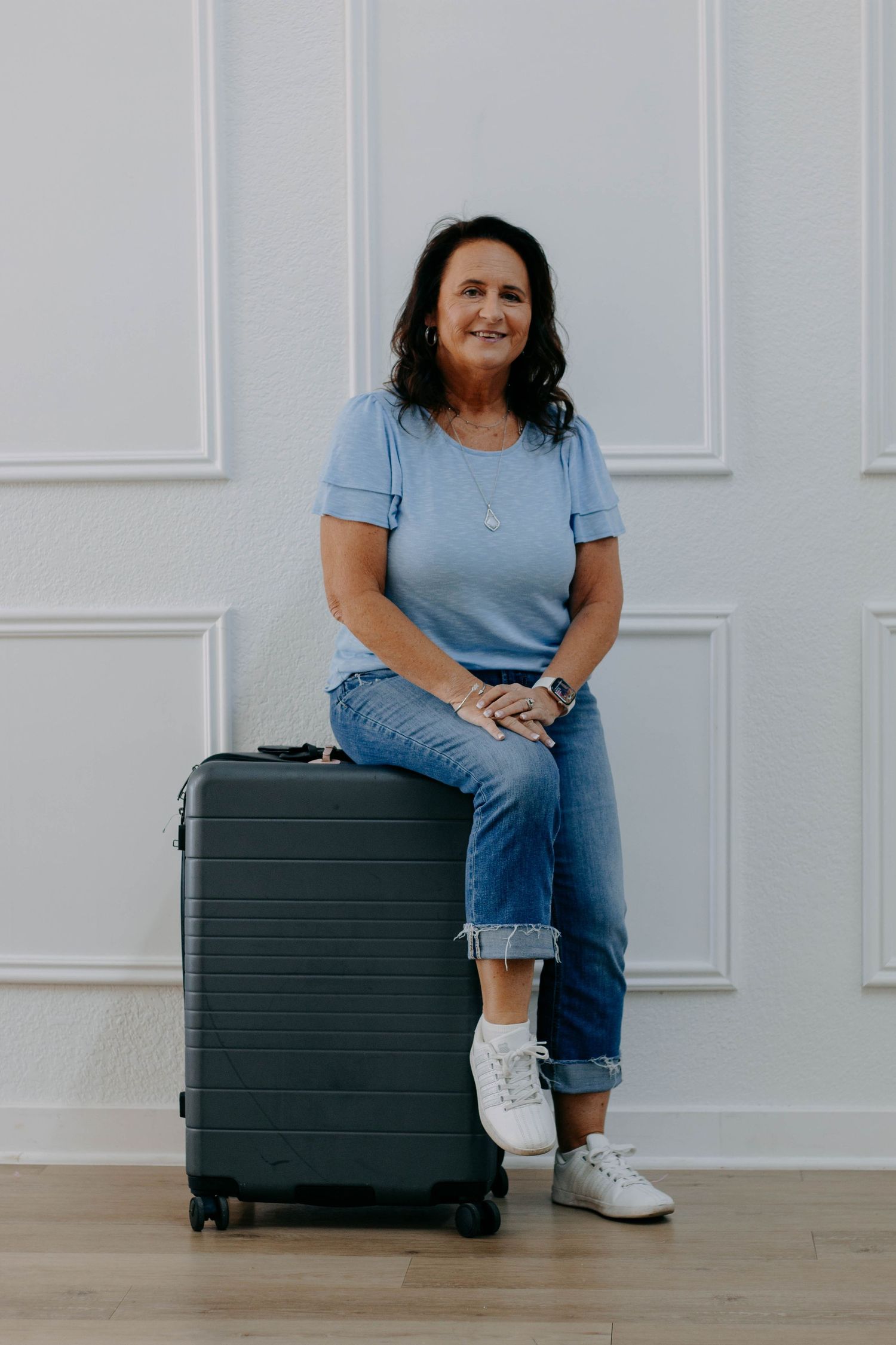 A middle-aged woman in a blue t-shirt and jeans sits on a dark carry-on suitcase against a white wall.