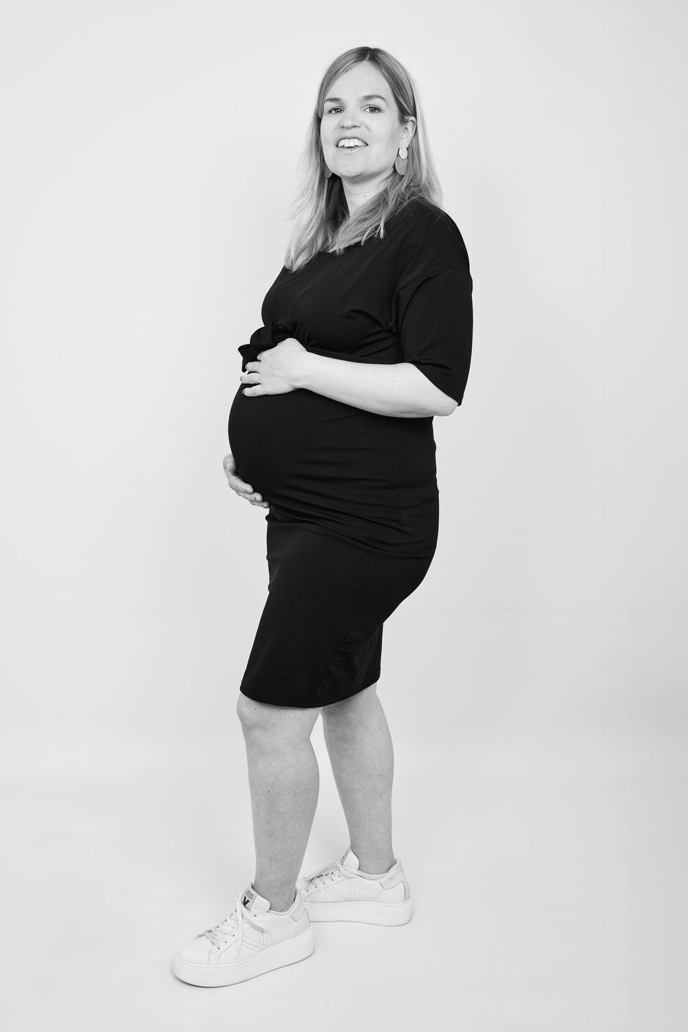 Smiling pregnant woman in black dress holding baby bump, standing against white background in black and white photo.