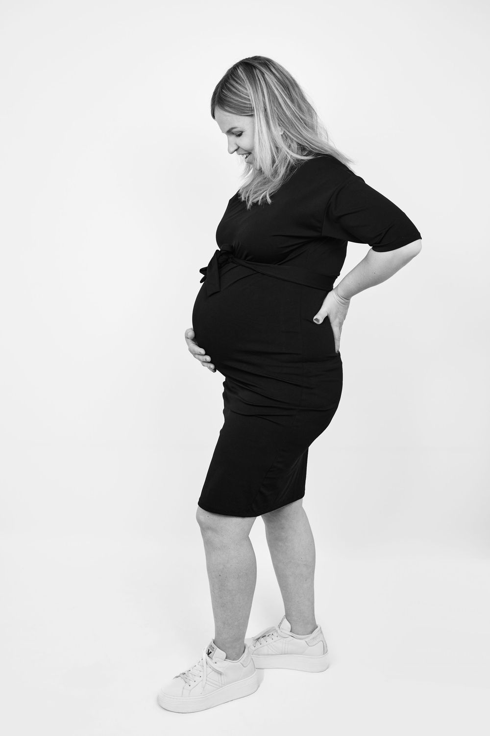 Smiling pregnant woman in black dress holding baby bump, black and white studio photo.