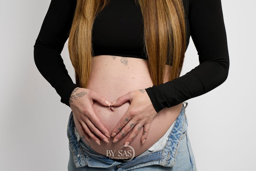 Pregnant woman in black crop top and jeans forming heart shape with hands on baby bump, maternity photo by SAS.