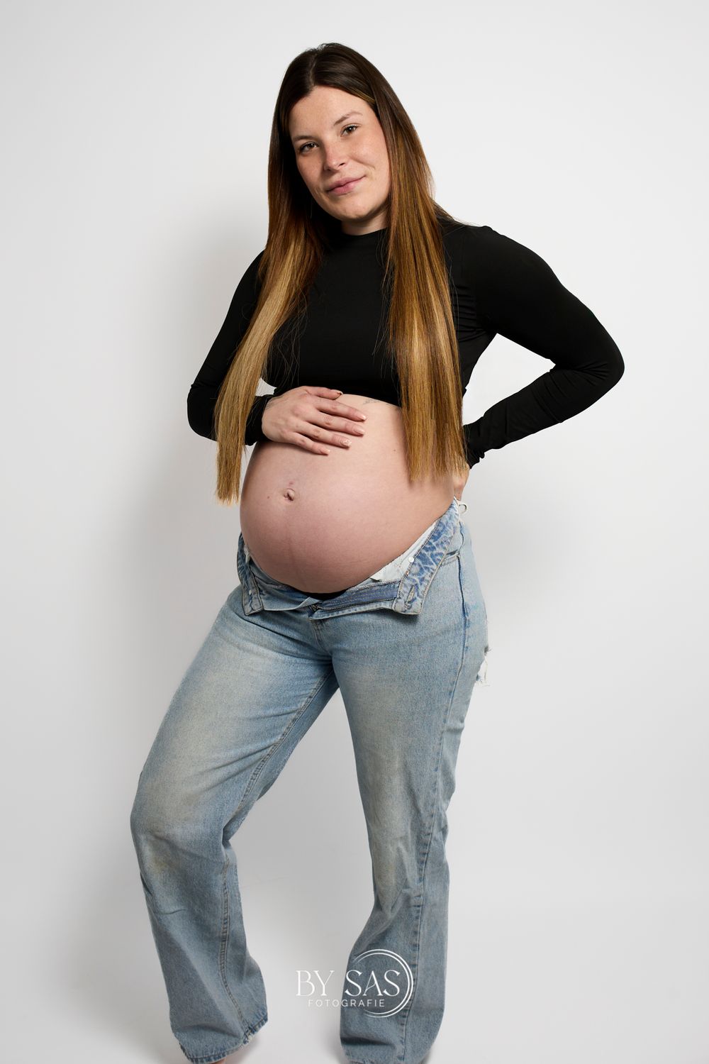 Pregnant woman in black turtleneck and open jeans cradling baby bump, posing for maternity photo shoot.