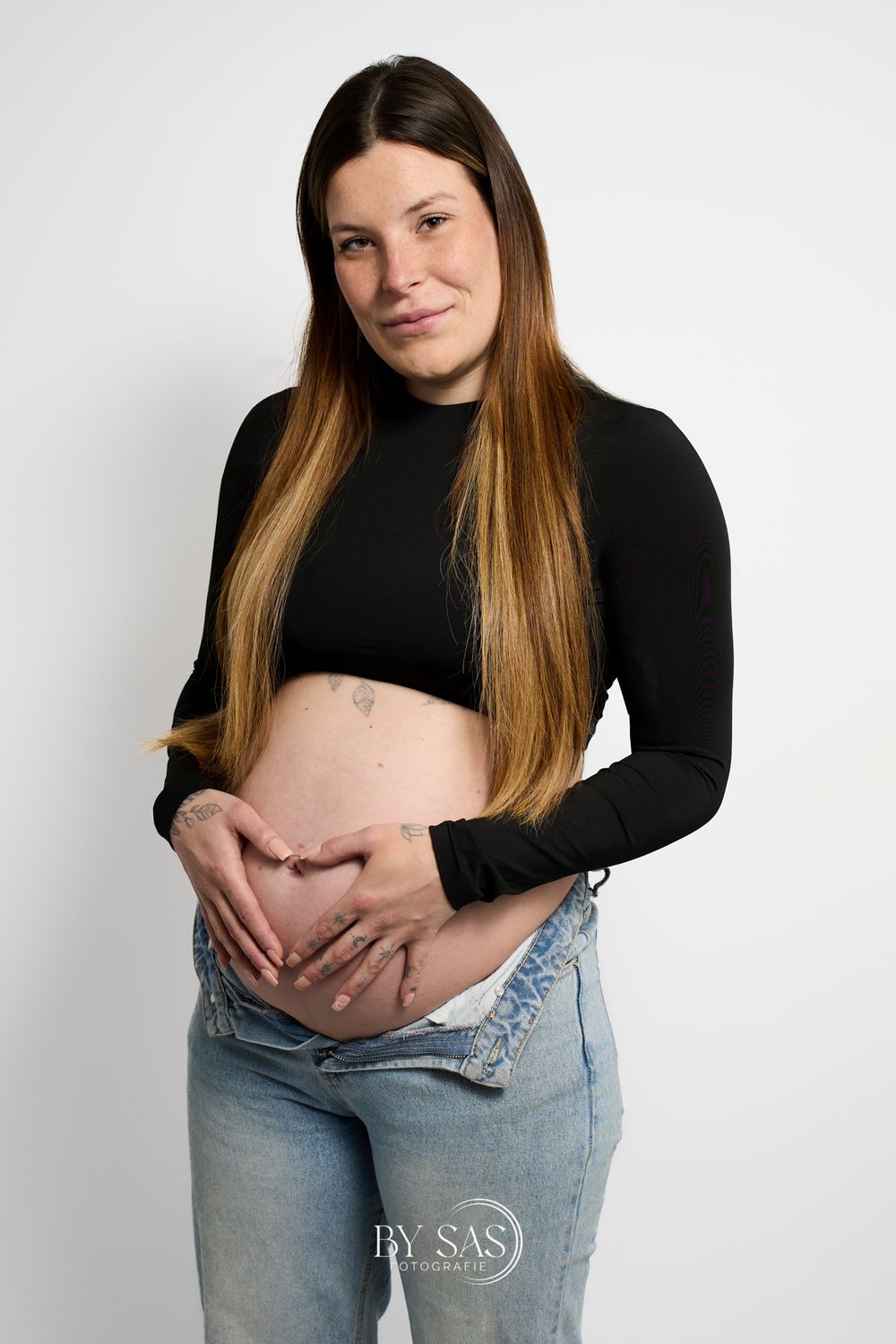 Pregnant woman in black crop top and jeans cradling baby bump, maternity photography studio portrait.