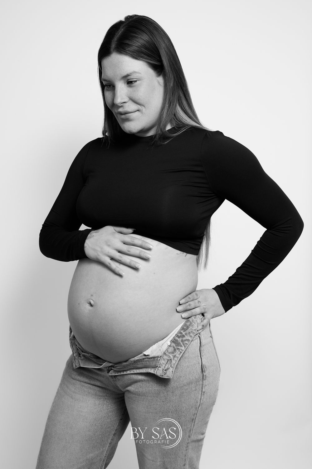 Black and white maternity photo of a pregnant woman in black top and jeans, cradling her baby bump.