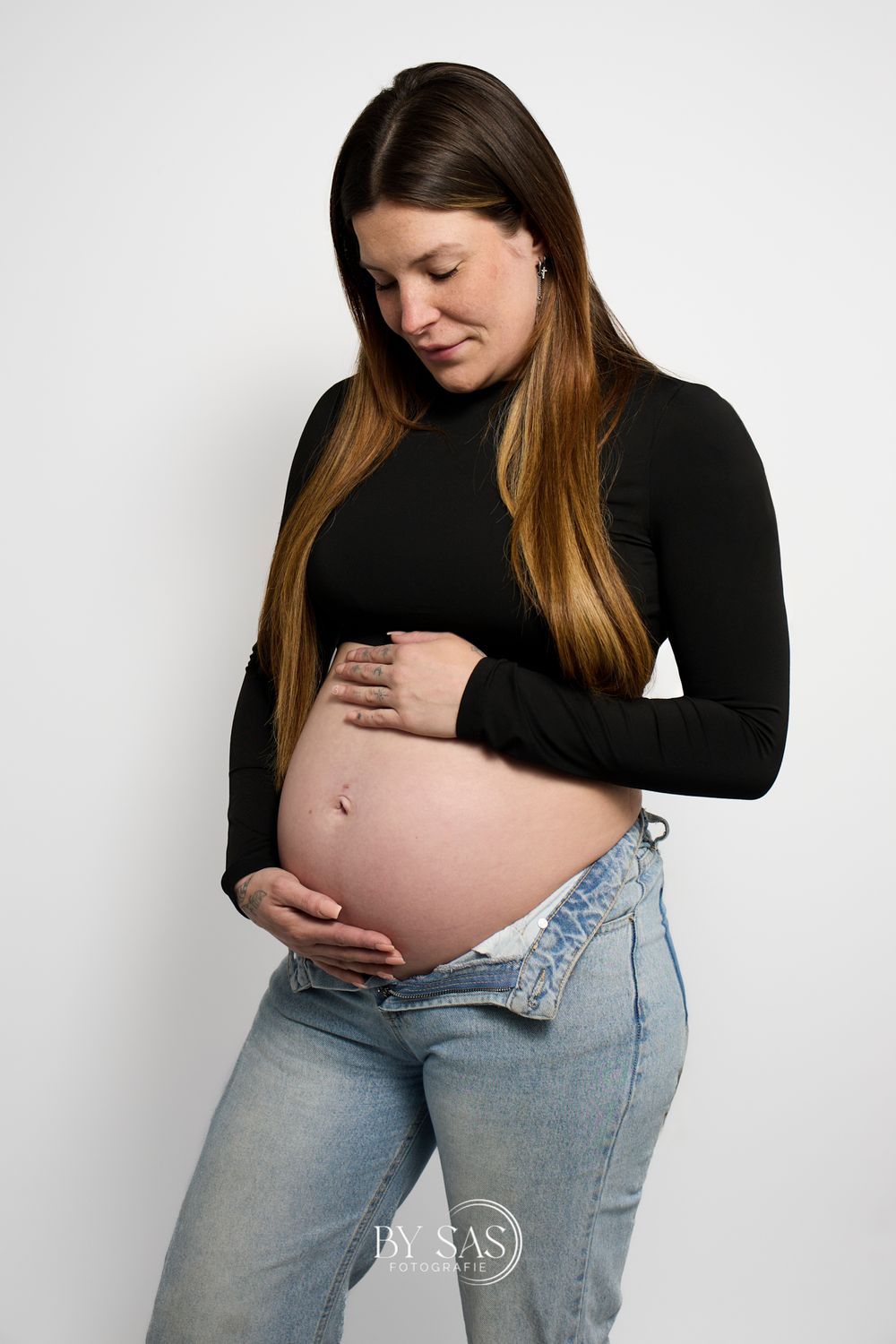 Pregnant woman in black top and open jeans cradling her bare baby bump against a white background.