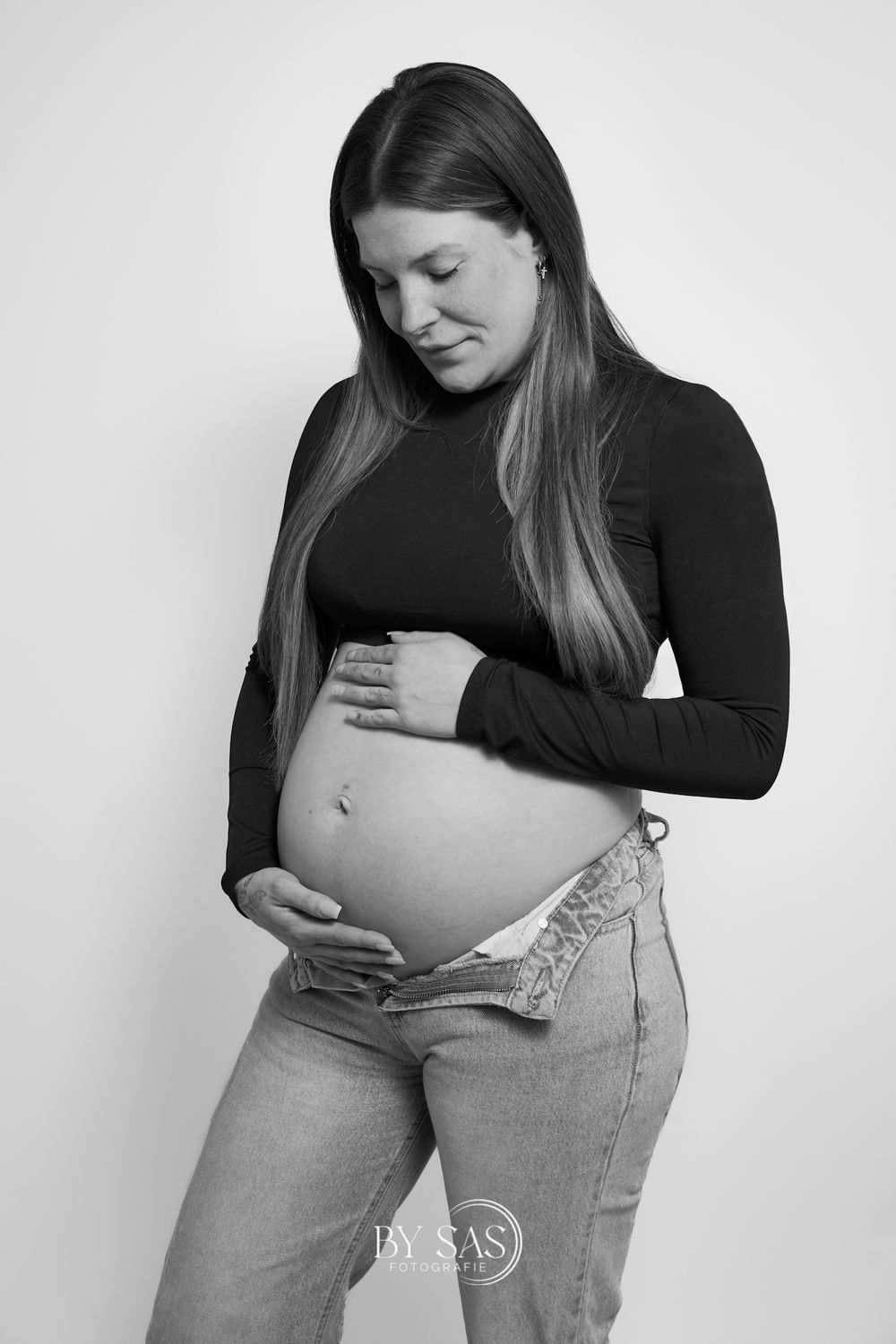 Black and white maternity photo of a pregnant woman in jeans cradling her baby bump in a studio.