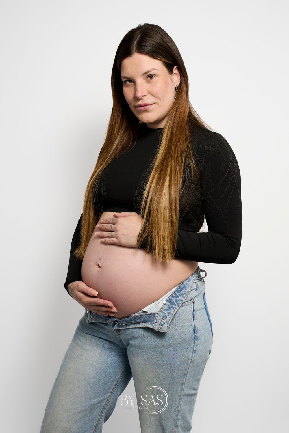 Pregnant woman in black crop top and open jeans cradling baby bump during professional maternity photo session.