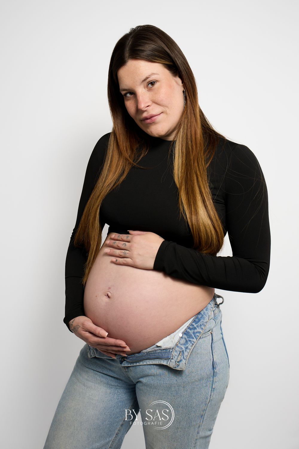 Pregnant woman in black turtleneck and jeans cradling her baby bump against a white background.