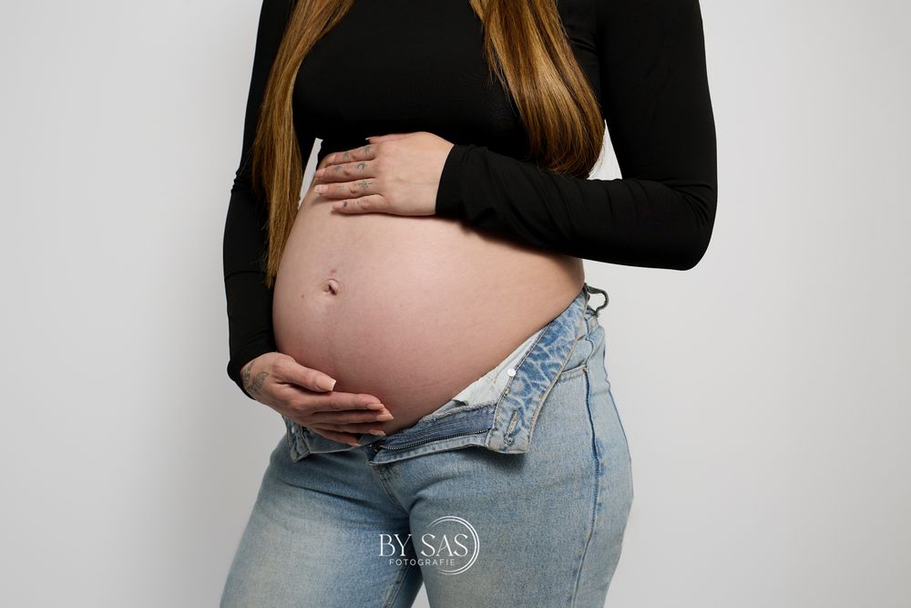 Pregnant woman in black top and unbuttoned jeans cradling her bare baby bump against a white background.