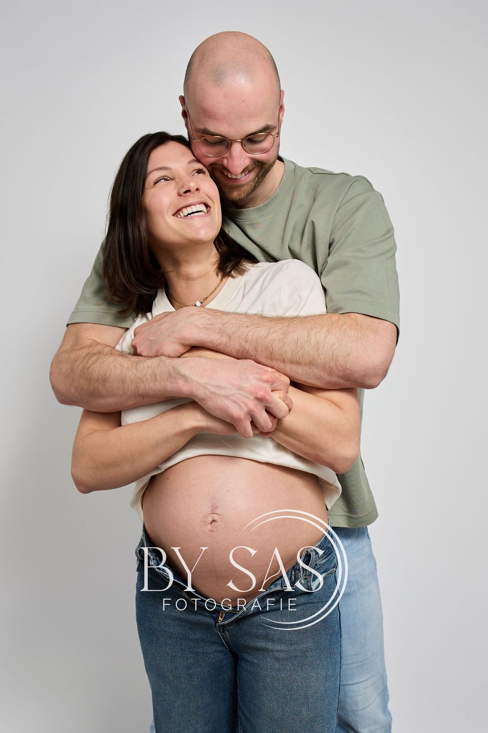 Pregnant woman smiling as her partner embraces her from behind, cradling her baby bump in a maternity photo shoot.