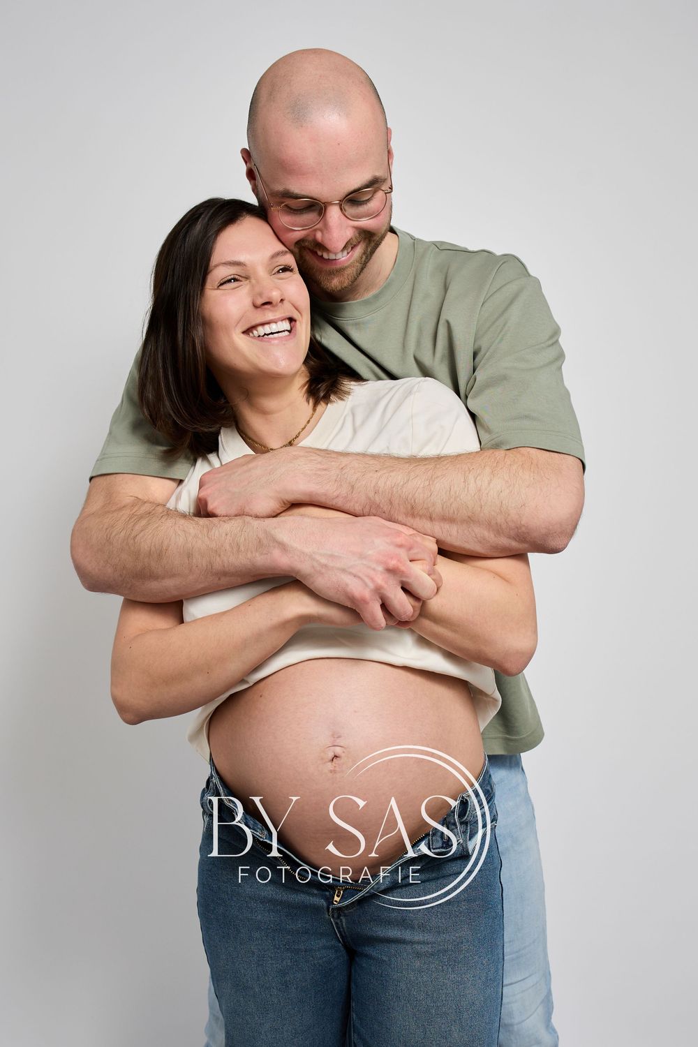Smiling pregnant woman embraced from behind by bald man with glasses in a maternity photography studio session.