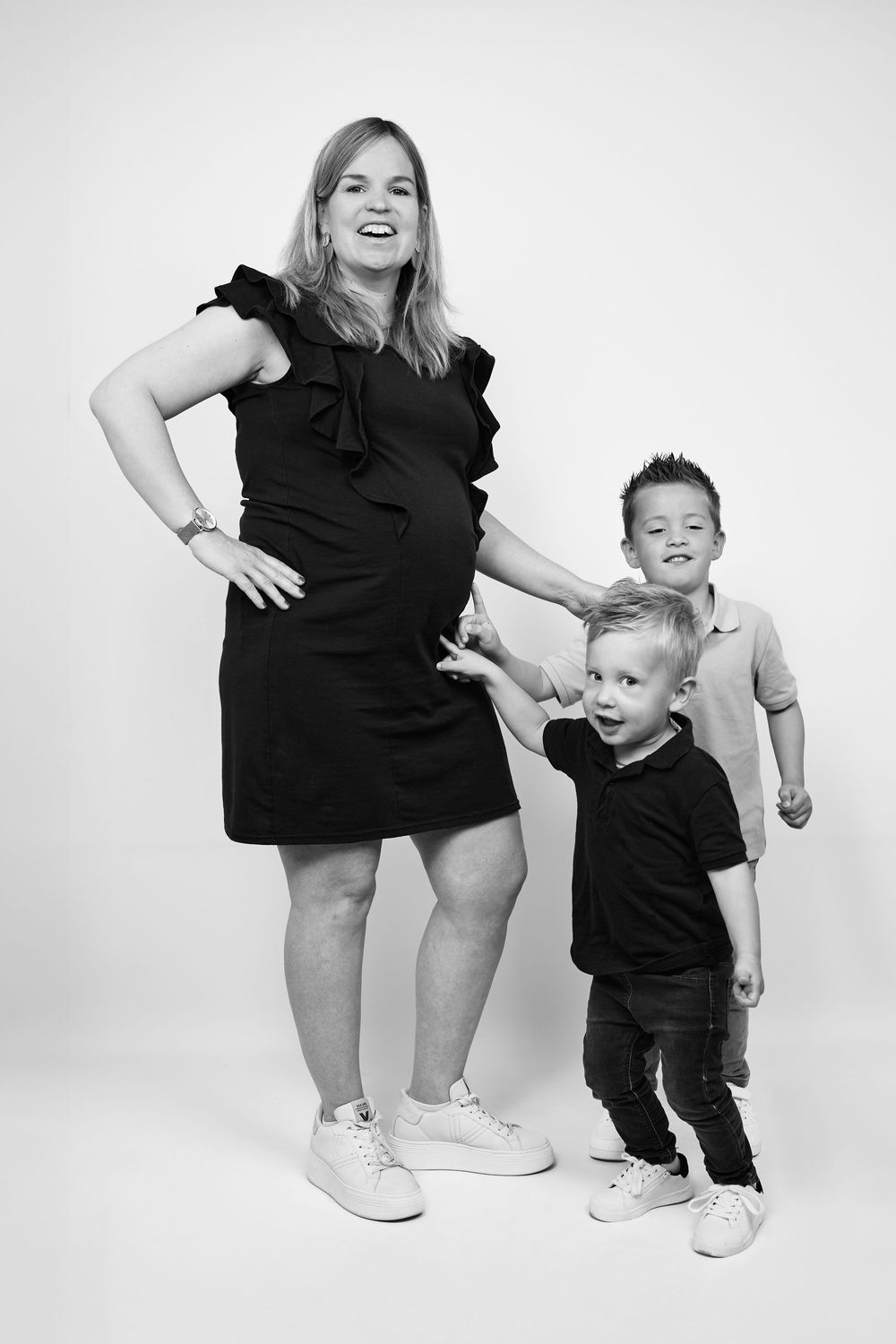 Black and white photo of a pregnant woman in a black dress smiling with two young boys in a studio setting.