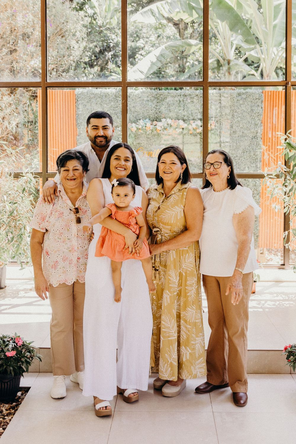 Multi-generational family portrait with six members smiling indoors, featuring a woman holding a baby in an orange outfit.