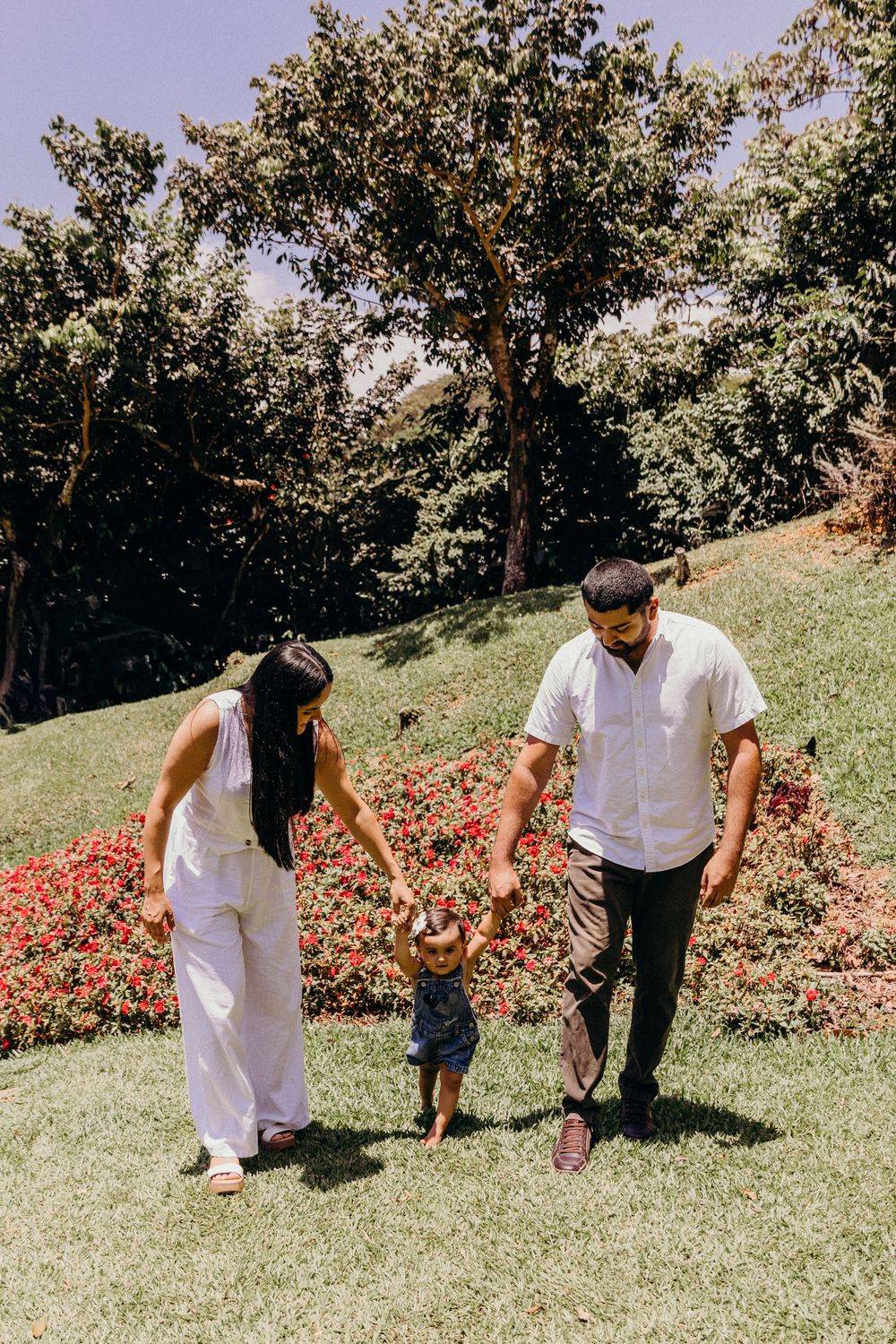 A young family walks hand-in-hand with their toddler on a sunny green lawn with red flowers and trees in the background.