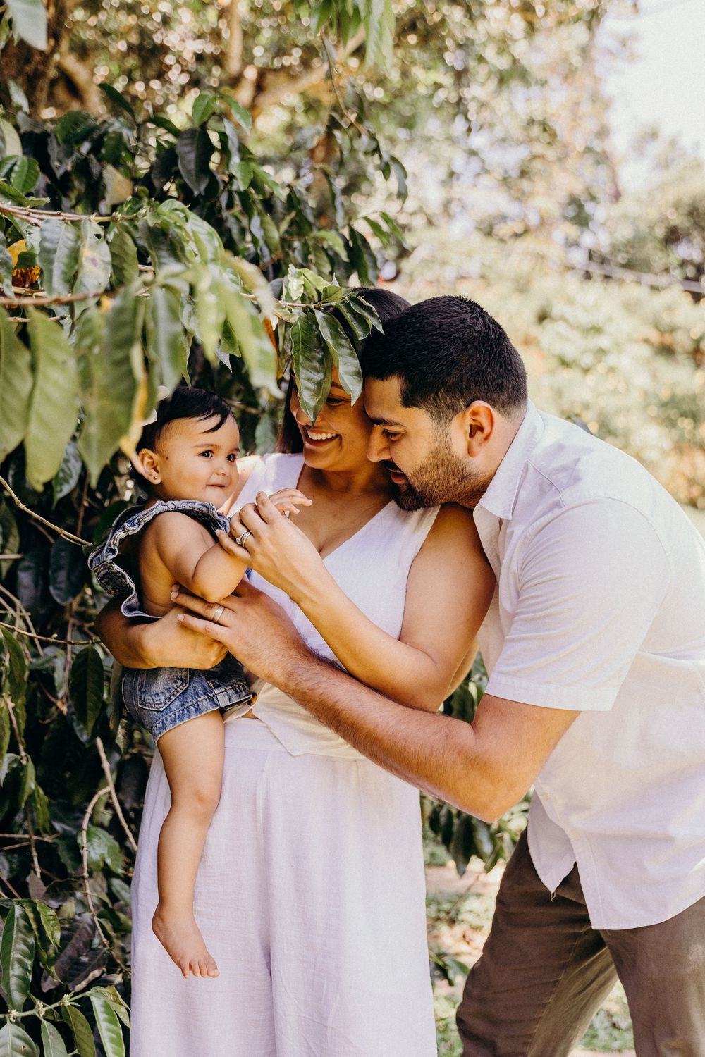 A happy young family with a baby girl poses outdoors under lush green trees in a warm, sunlit garden setting.