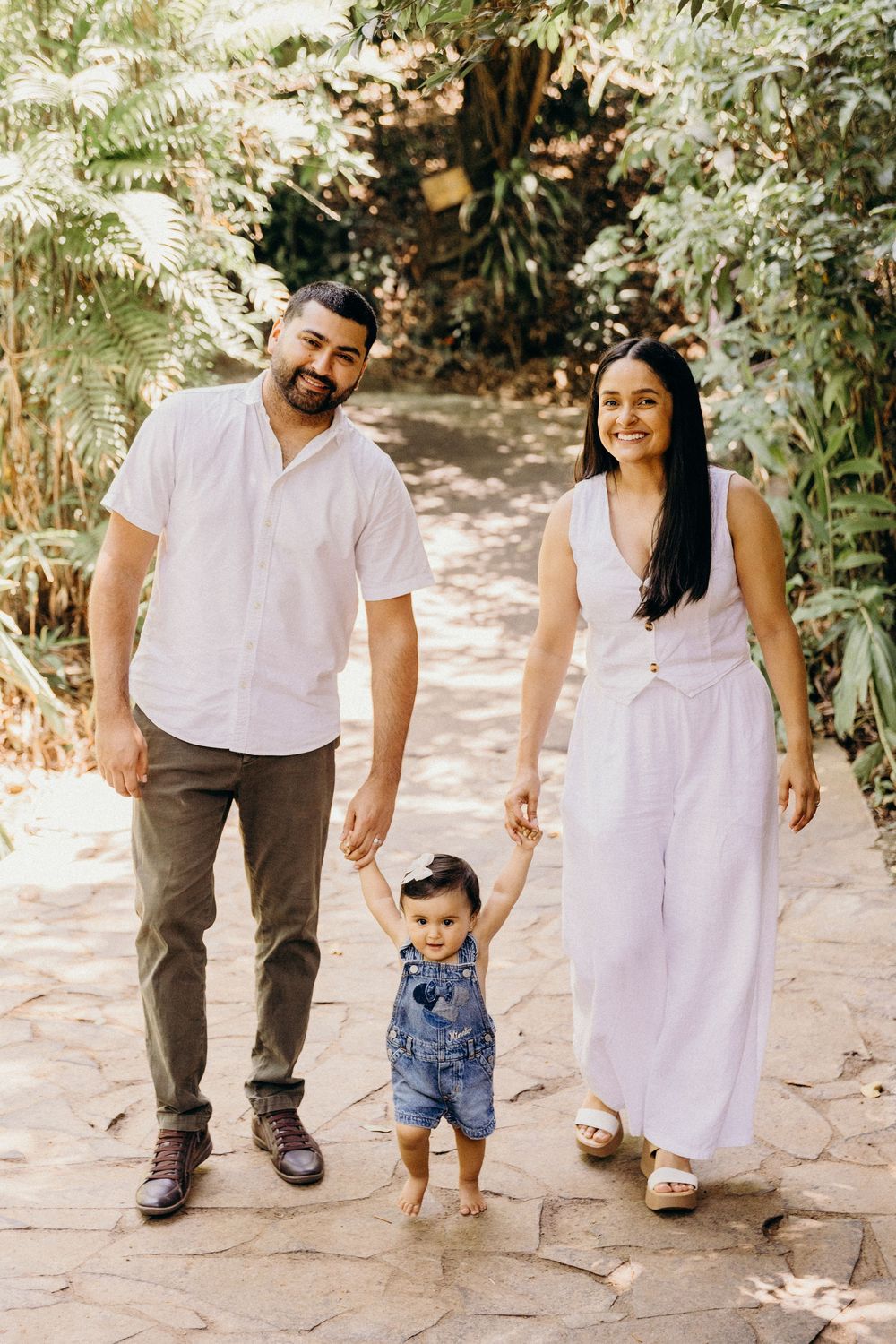 Happy family with parents holding hands of baby toddler walking on stone path in lush green garden.