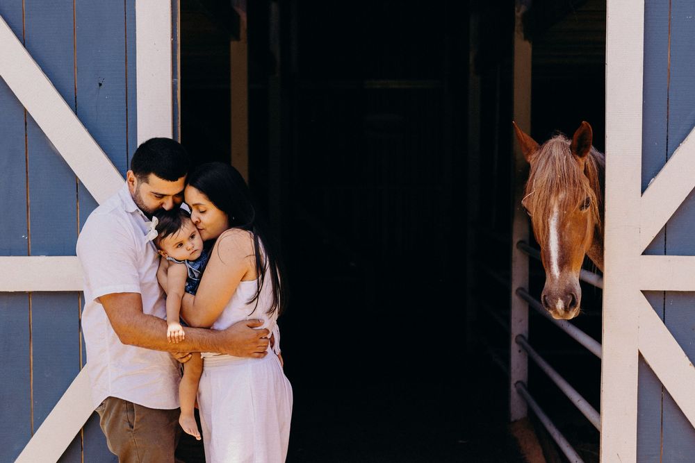 Family embracing near a blue barn door with a brown horse peeking out behind them.