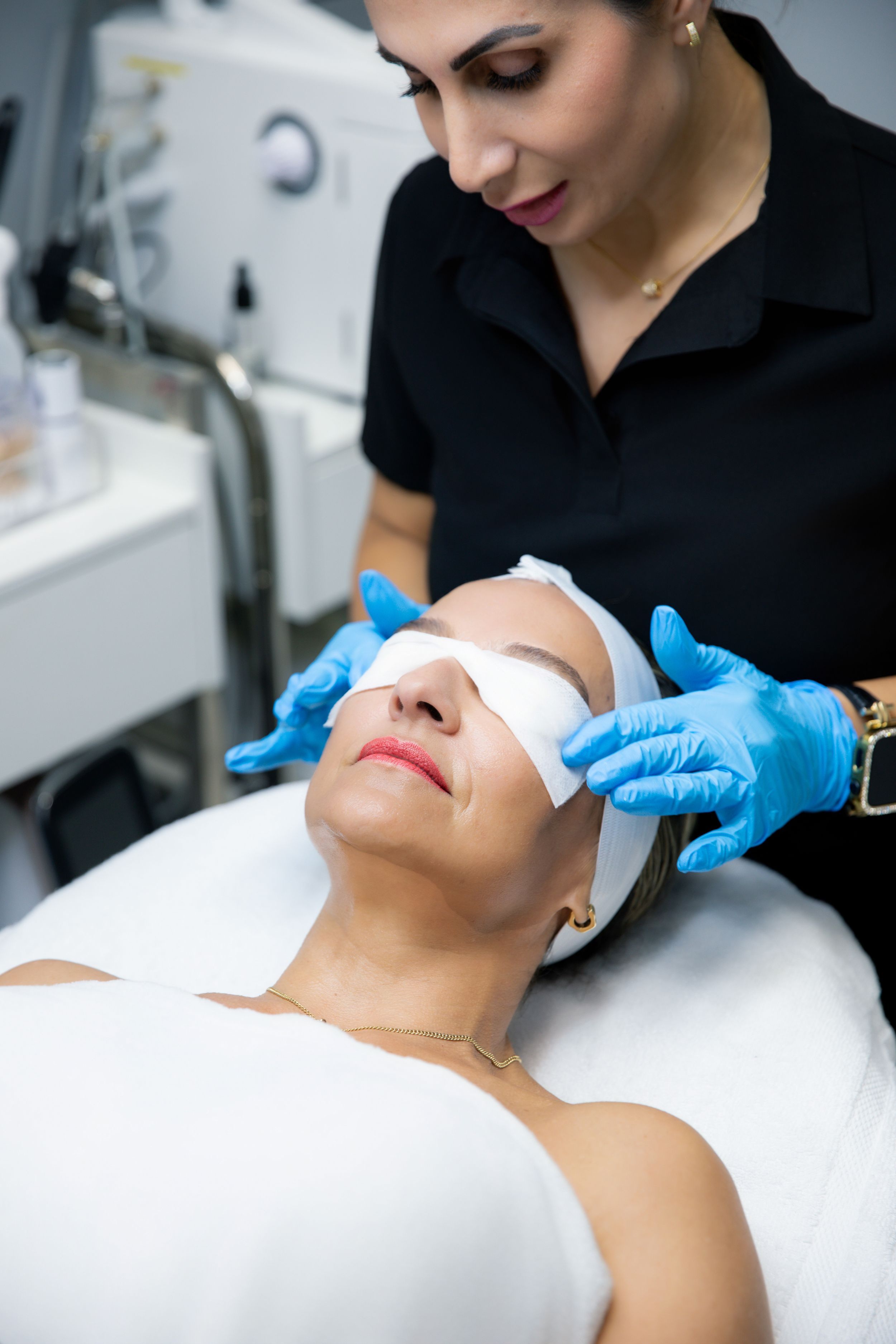 Esthetician in blue gloves applying facial treatment to relaxed client lying on spa bed in modern beauty salon.