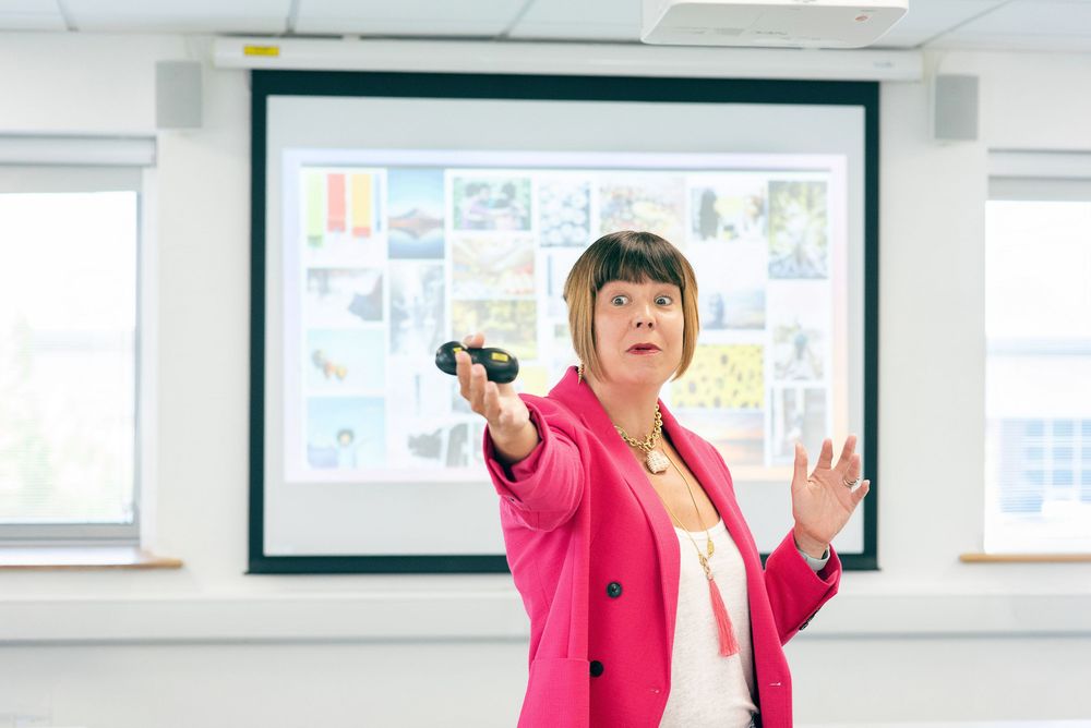 Someone in a bright pink blazer gestures while giving a presentation in a classroom setting with a projector screen behind.