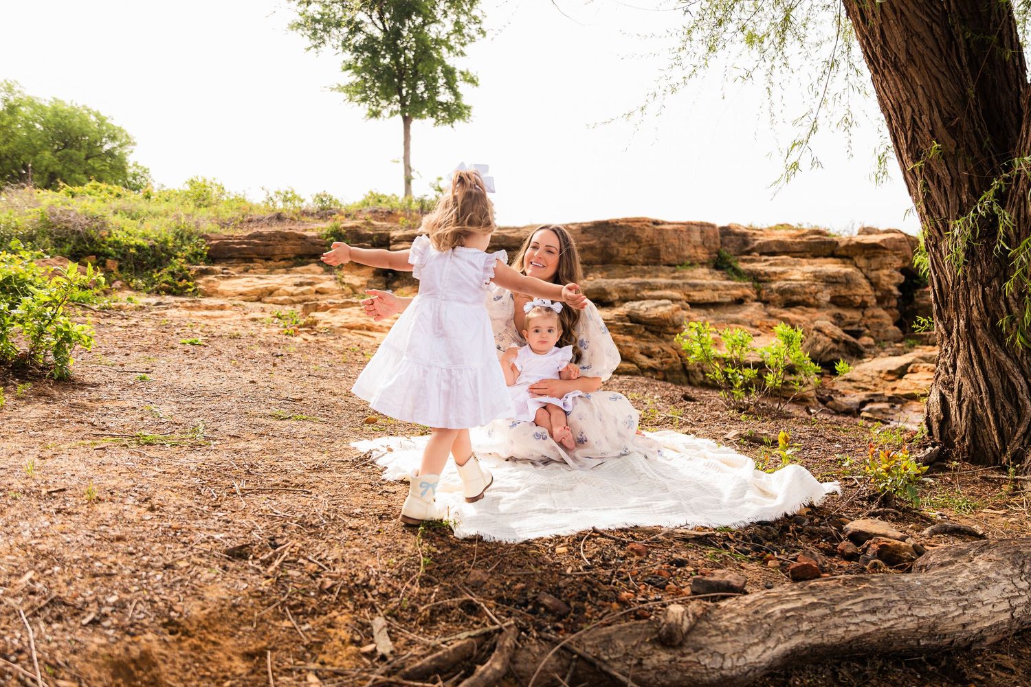 A mother in white sits with two young daughters in white dresses on a blanket under a tree outdoors.