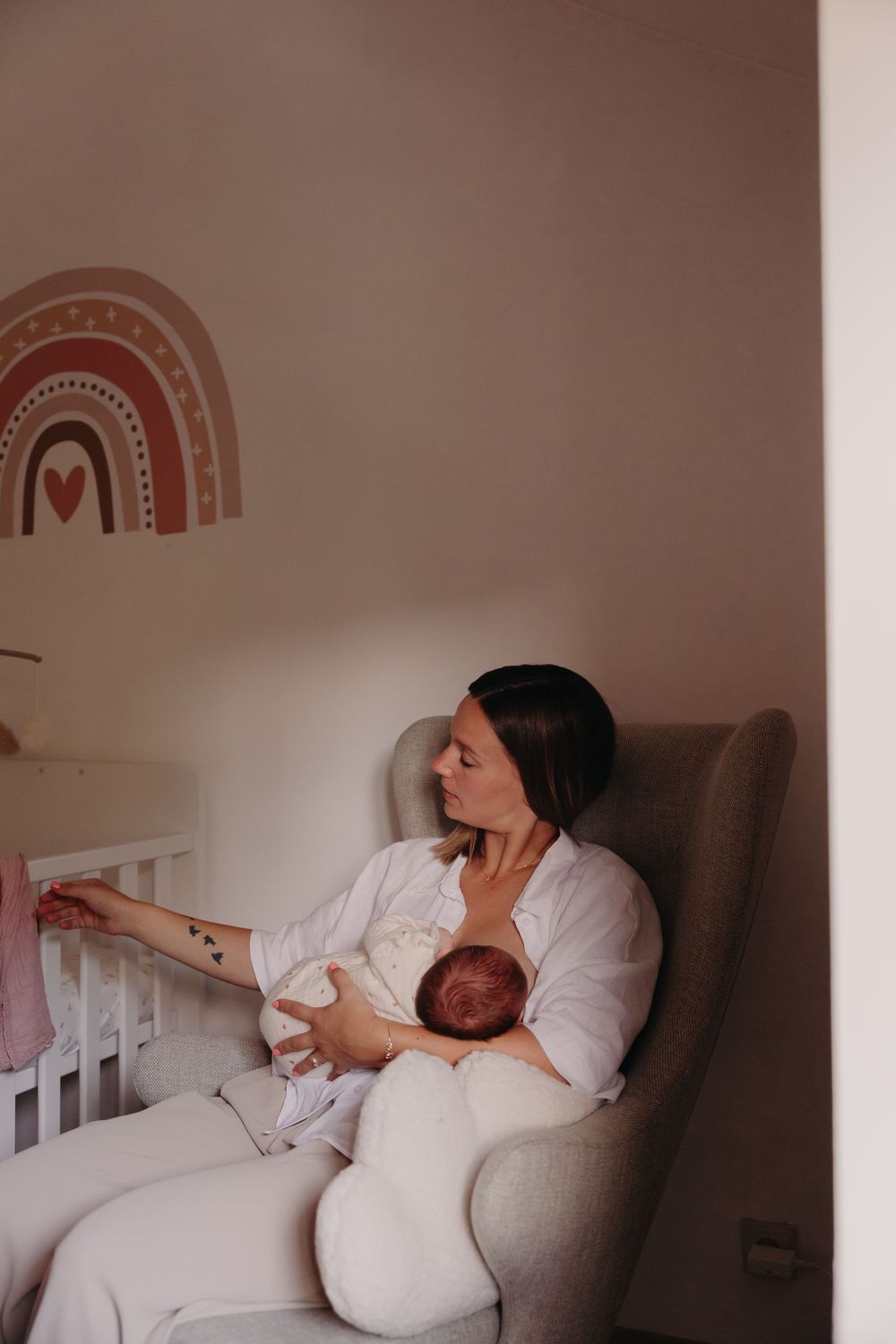 Mother sitting in nursery glider chair holding newborn baby beside crib with rainbow wall decal.