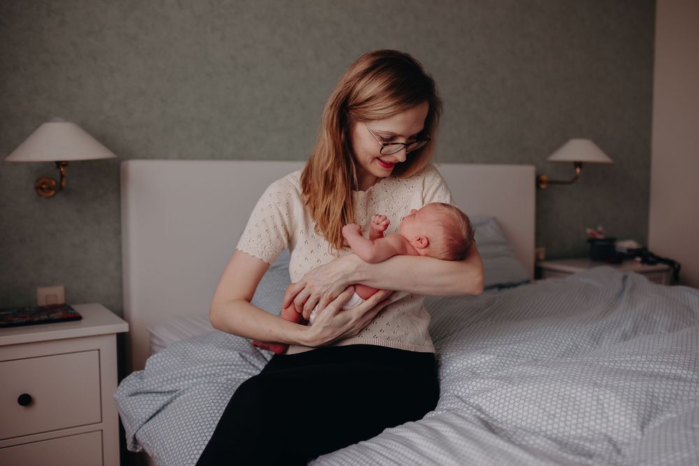 Young mother with glasses sitting on bed cradling her newborn baby in a cozy bedroom setting.