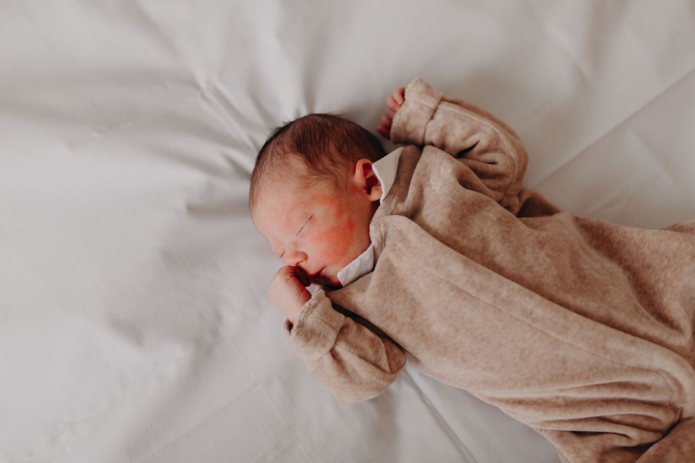 Newborn baby in beige sleepsuit lying peacefully on white bedding.