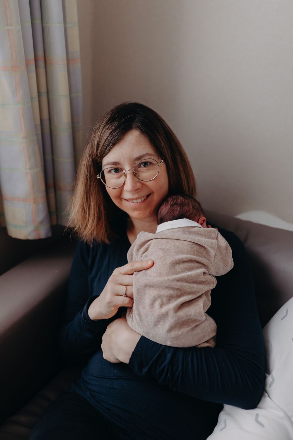Smiling mother with glasses holding swaddled newborn baby while sitting on sofa.