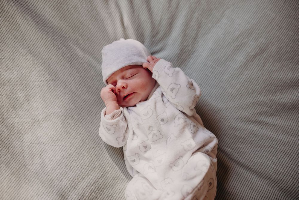 Peaceful sleeping newborn baby in white hat and patterned onesie lying on gray blanket.