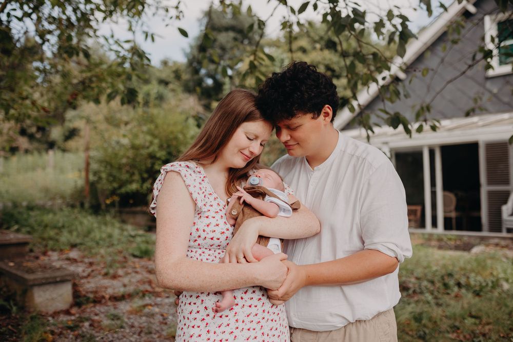 Couple tenderly holding newborn baby outdoors in garden setting near rustic home.