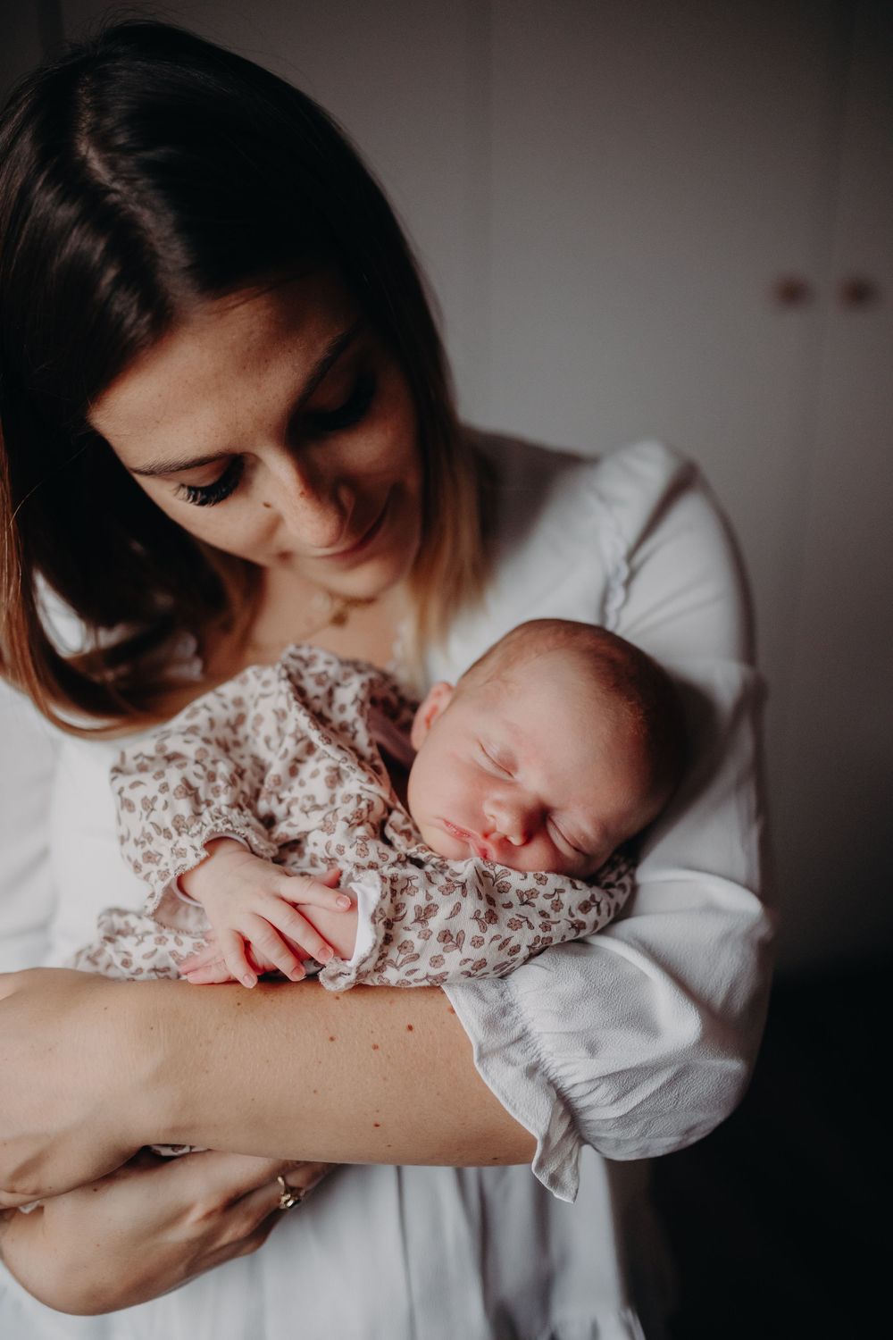A smiling mother in a white blouse tenderly holding her sleeping newborn baby.