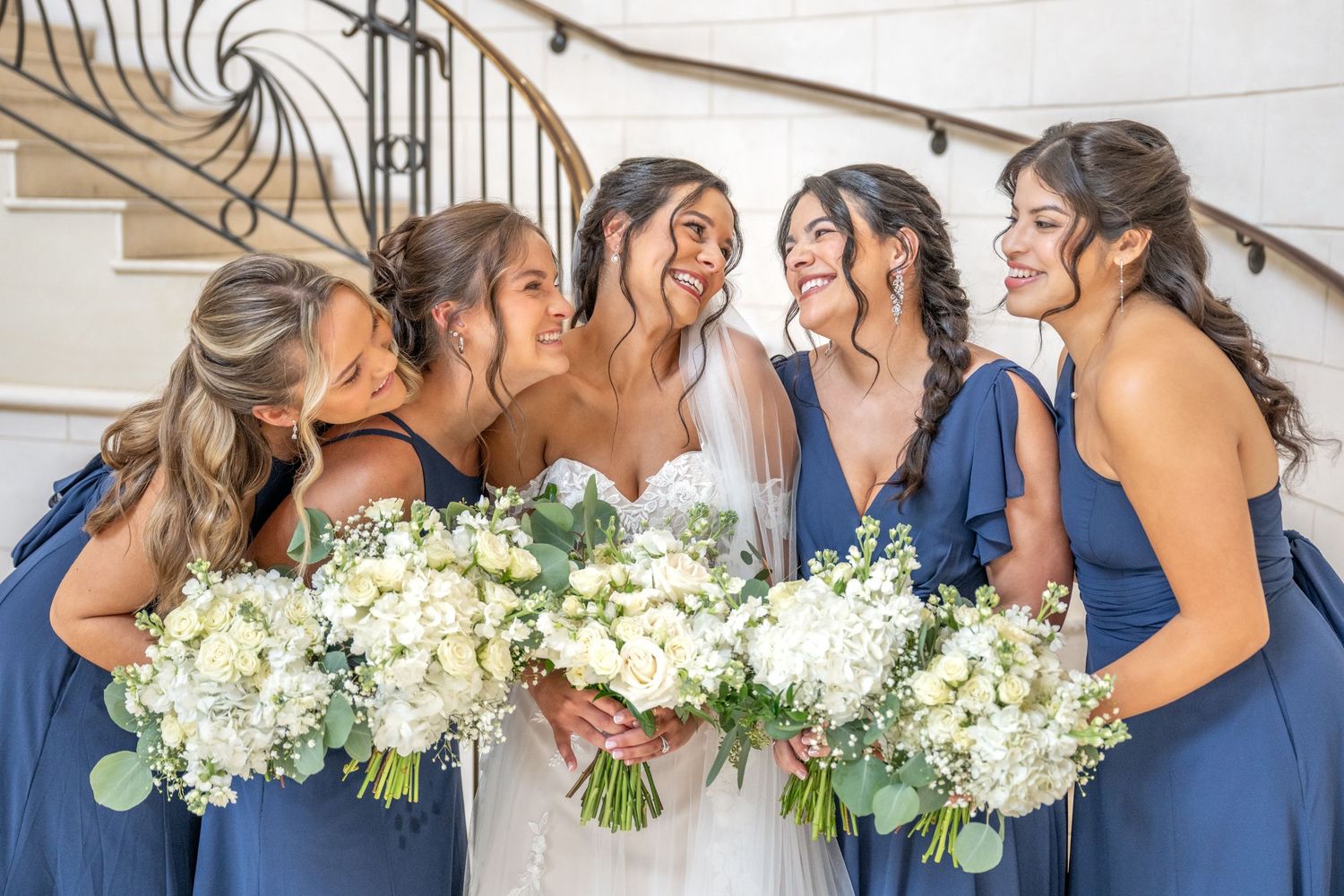 Bride in white gown with bridesmaids in blue dresses holding white floral bouquets, laughing on staircase.