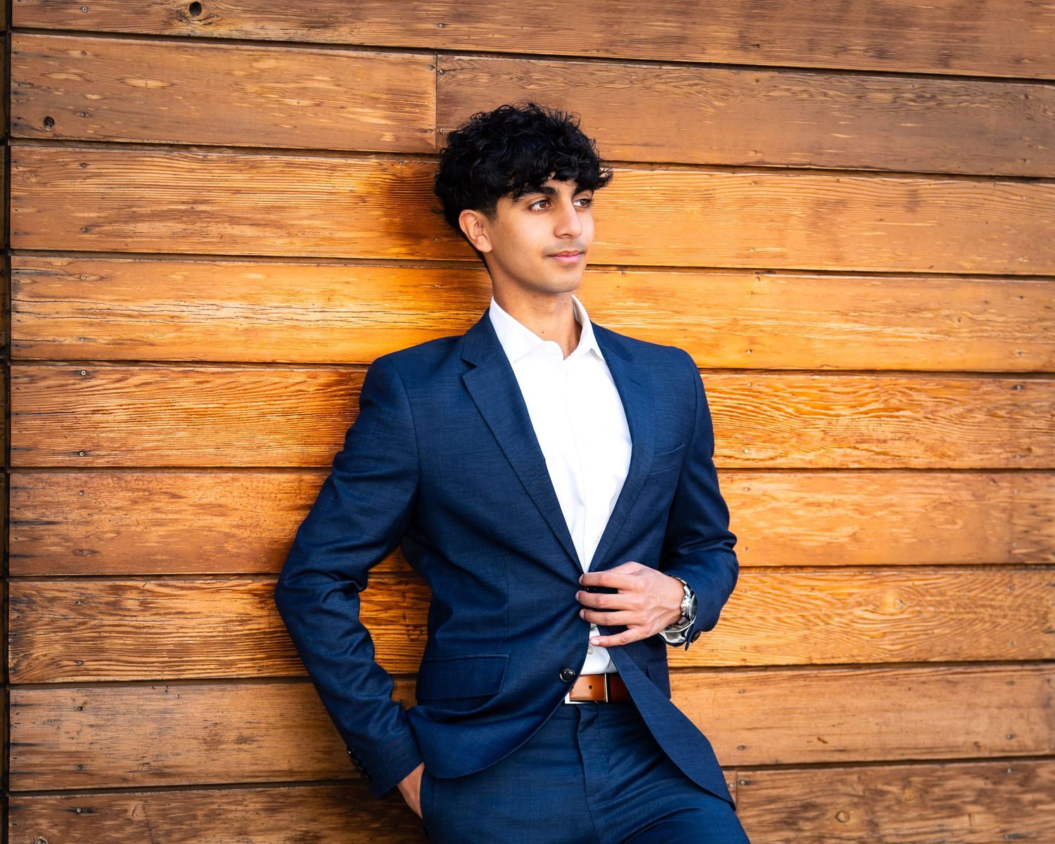 Young man in navy blue suit and white shirt posing confidently outdoors near stone column with green foliage background.
