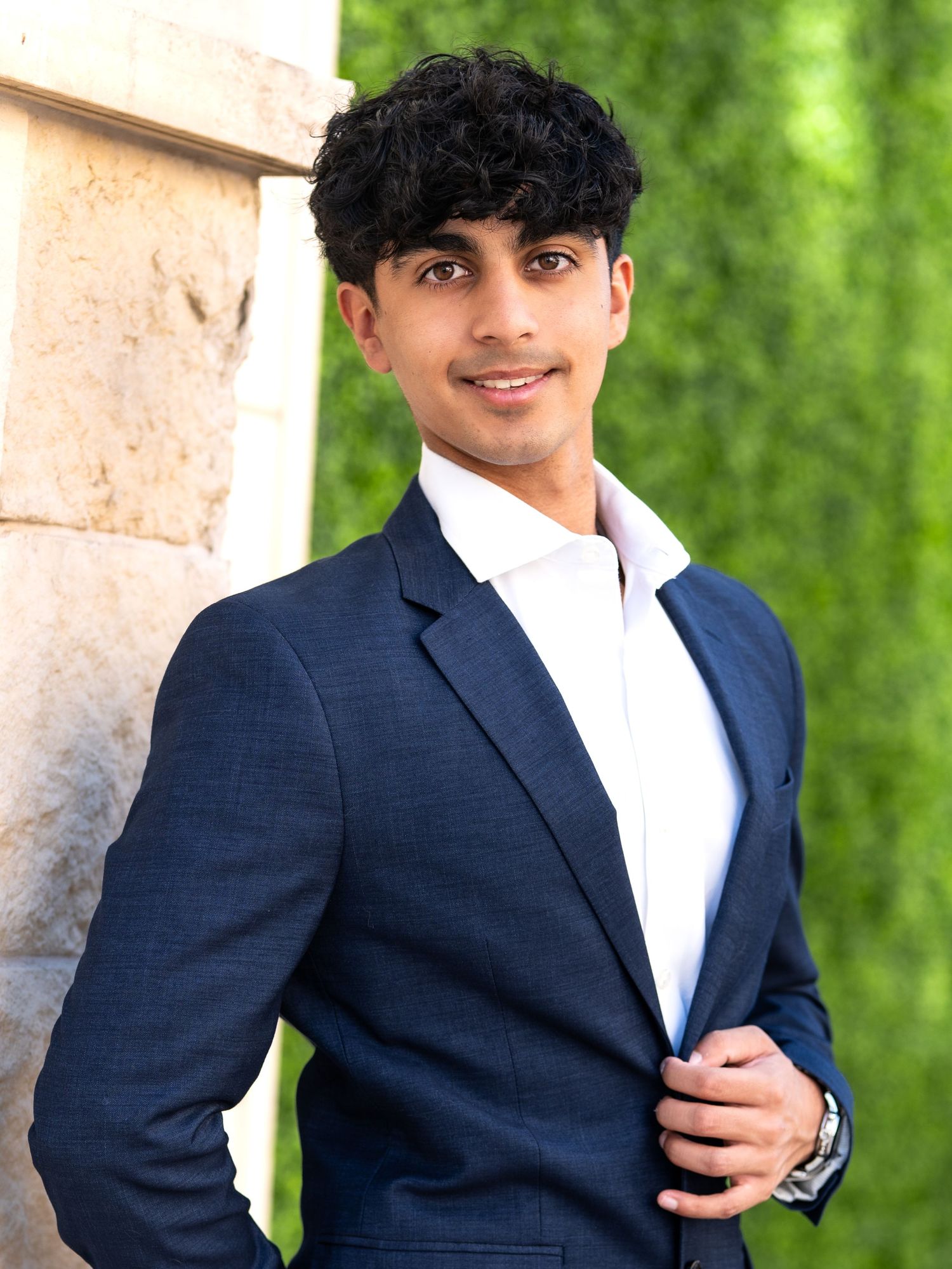 Young man in navy blue suit and white shirt posing confidently outdoors near stone column with green foliage background.
