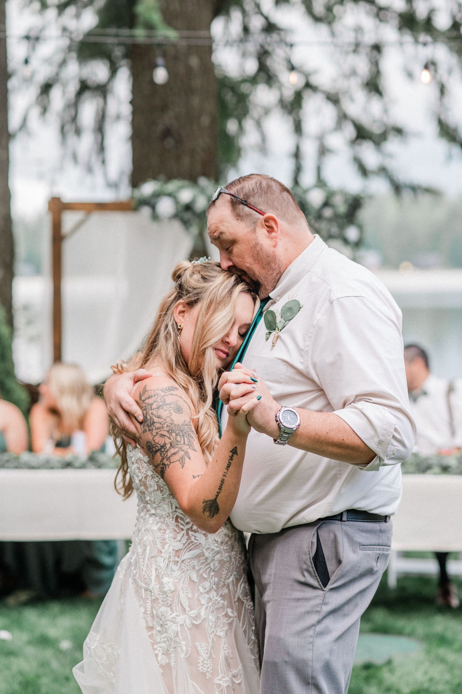 Emotional father-daughter wedding dance outdoors, bride in lace gown with tattoos, tearful embrace under string lights.
