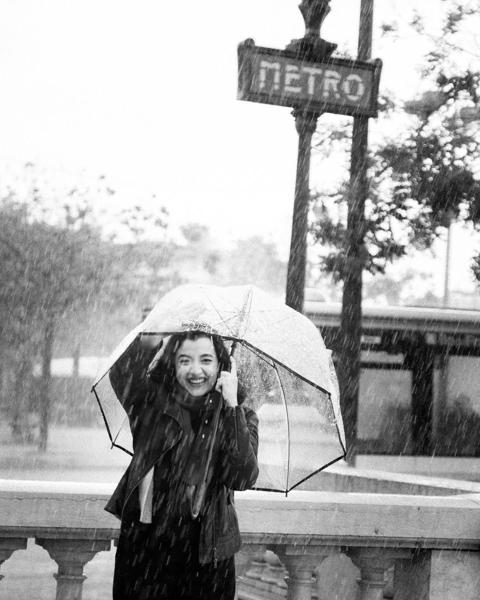 Black and white photo of a smiling woman holding a clear umbrella in the rain near a Paris Metro sign.
