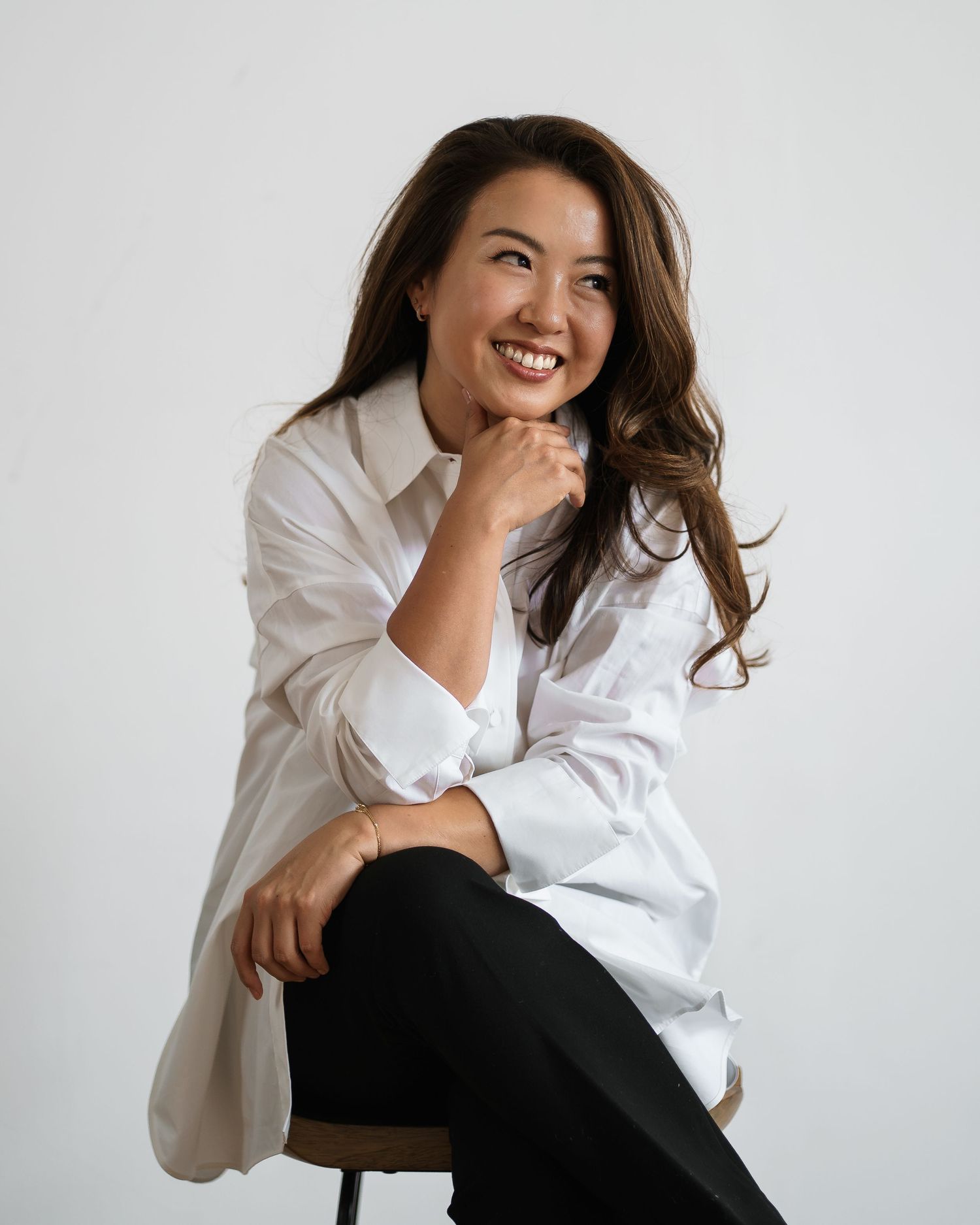 Smiling woman in white shirt and black pants sitting on stool, posing confidently against white background.