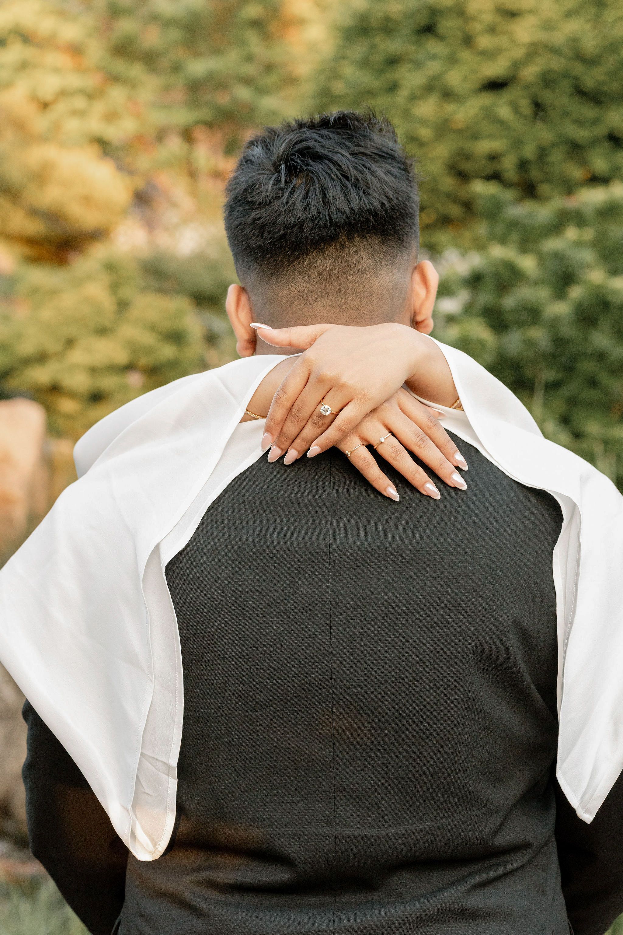 Bride with engagement ring embracing groom from behind in a garden setting during wedding celebration.