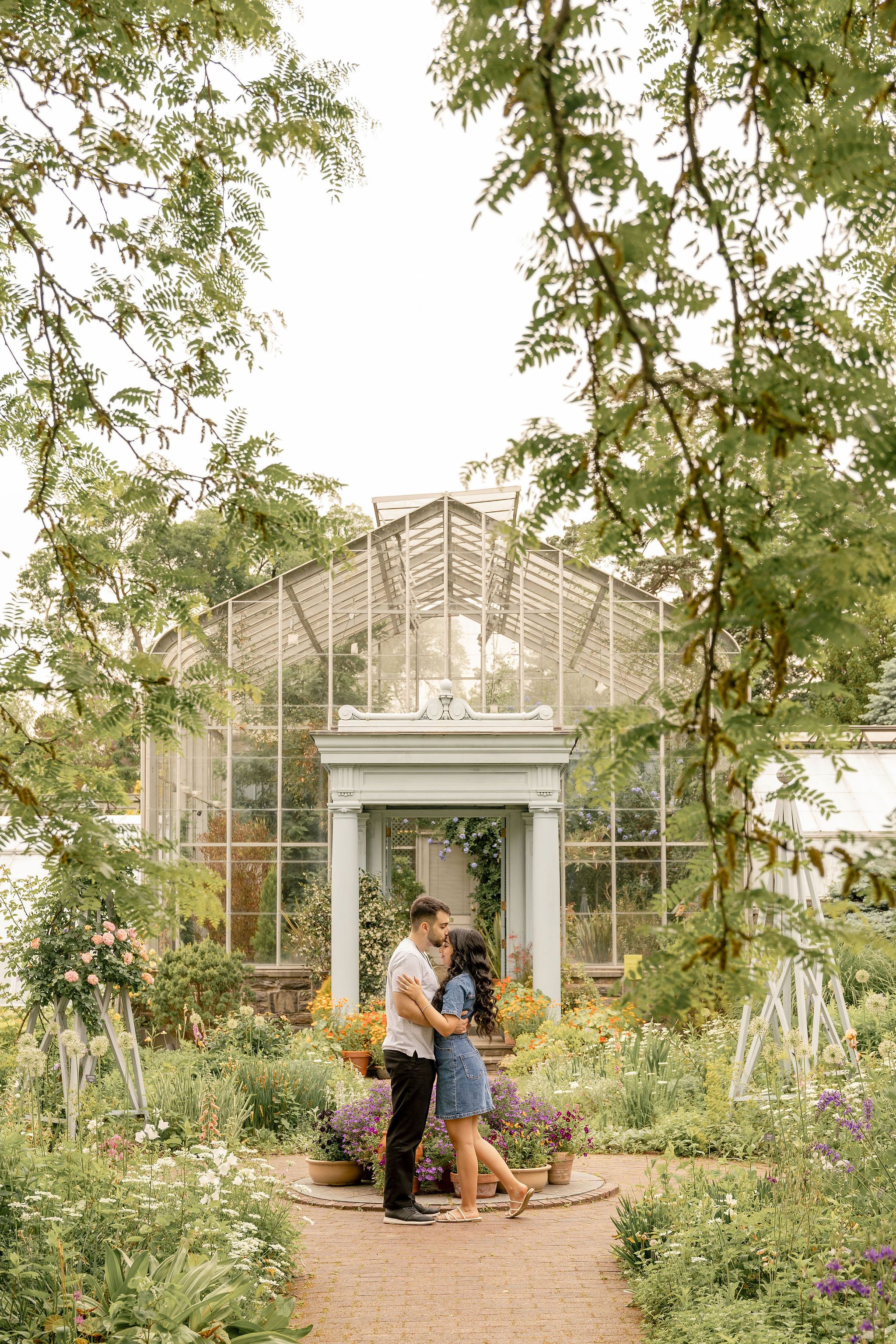 Couple sharing a romantic embrace in a lush garden with a charming greenhouse backdrop.