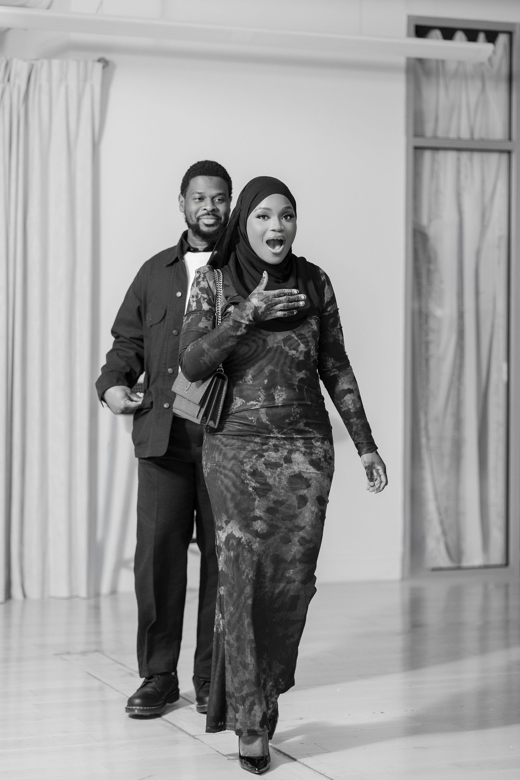 Black and white photo of a man and woman walking together, woman wearing hijab and floral dress, looking surprised.