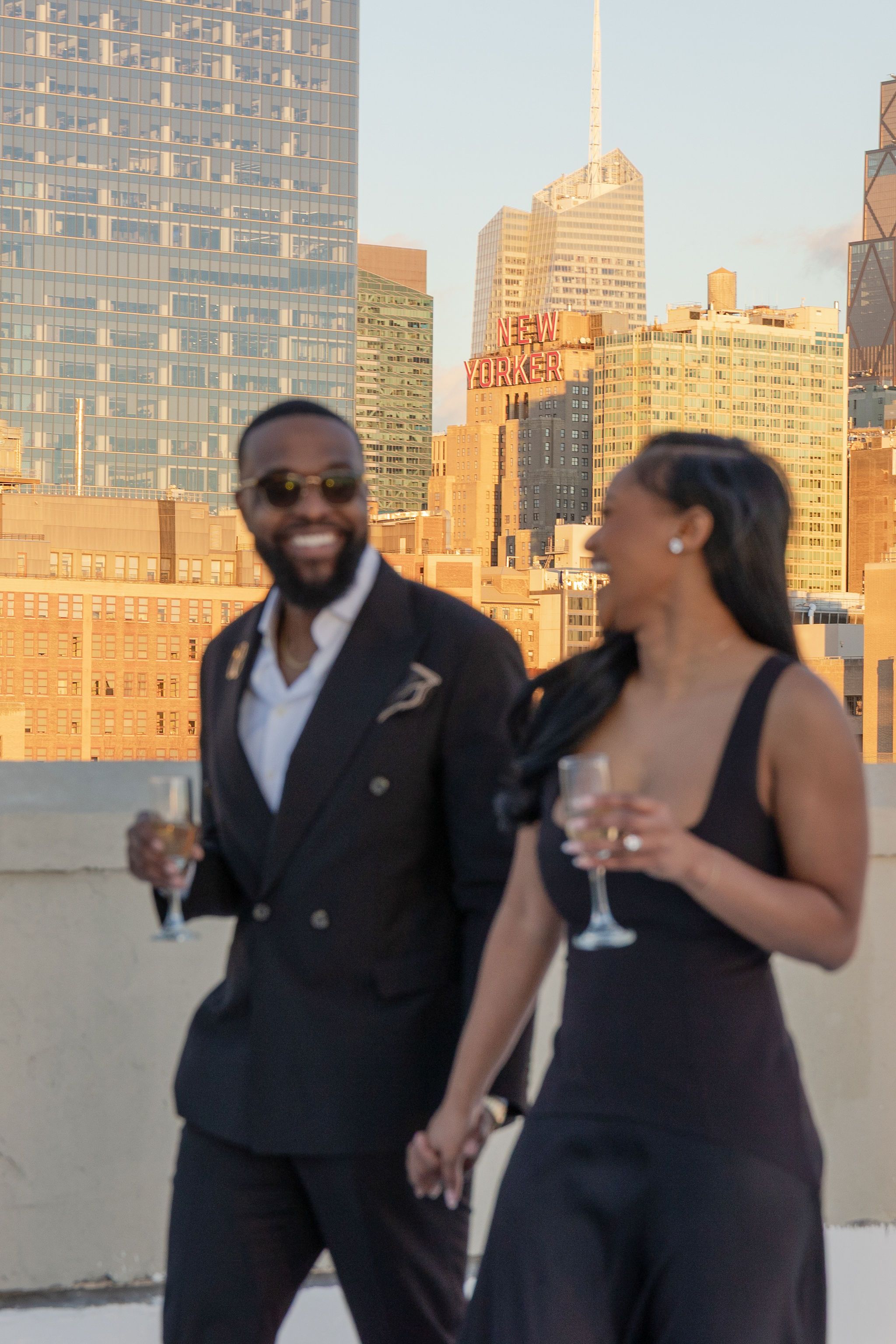 Elegant couple in formal wear holding champagne glasses on a rooftop with NYC skyline at golden hour.